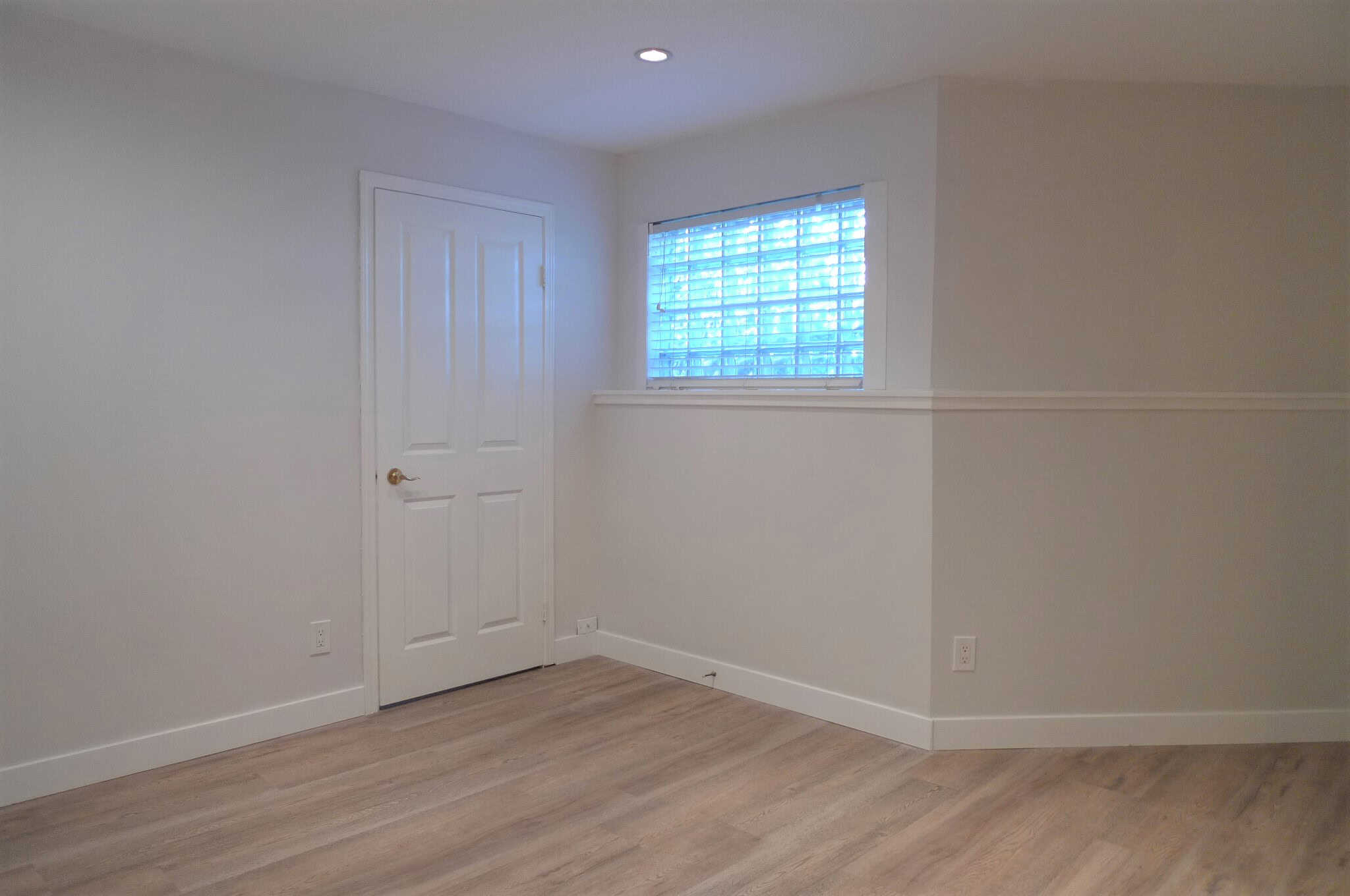 Bright, clean interior room with white walls, a window with blinds, and light wood flooring, perfect for a home or rental property.