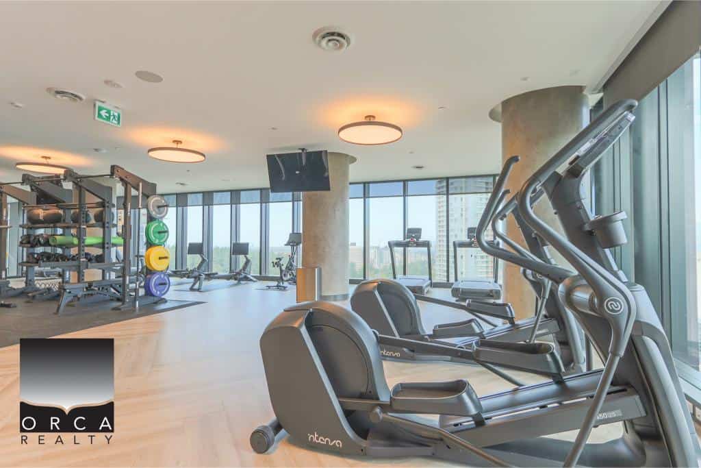 Modern gym facilities at Orca Realty Inc. featuring treadmills, elliptical machines, and weight training equipment in a bright, high-rise setting with floor-to-ceiling windows.