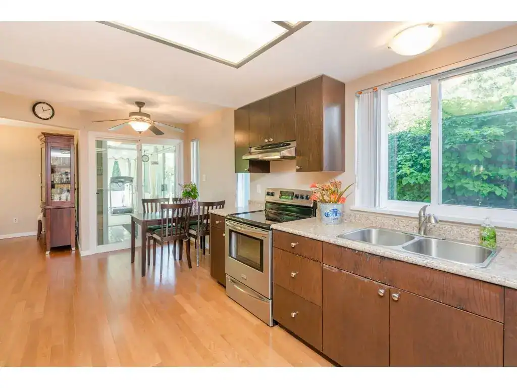 Bright kitchen with wooden cabinets, stainless steel stove, granite countertops, large window and view of lush greenery, open layout connects to dining area, ideal for family meals and entertaining.