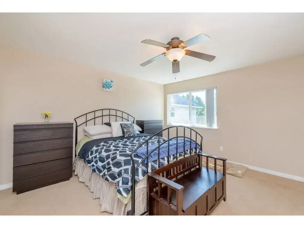 Bright bedroom with queen-size bed, ceiling fan, dark wood dresser, and natural light from large window, ideal for Vancouver Island homes.