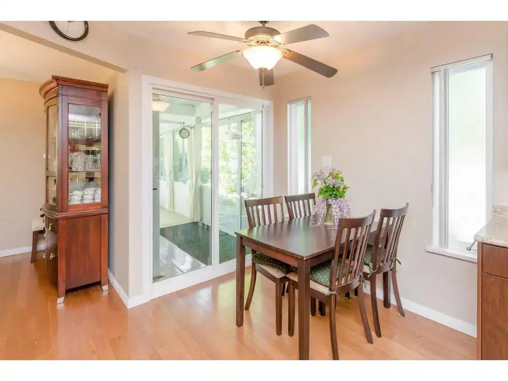 Bright dining room with wood flooring, sliding glass doors leading to a sunroom, and a modern ceiling fan, ideal for family meals and entertaining in BC homes.