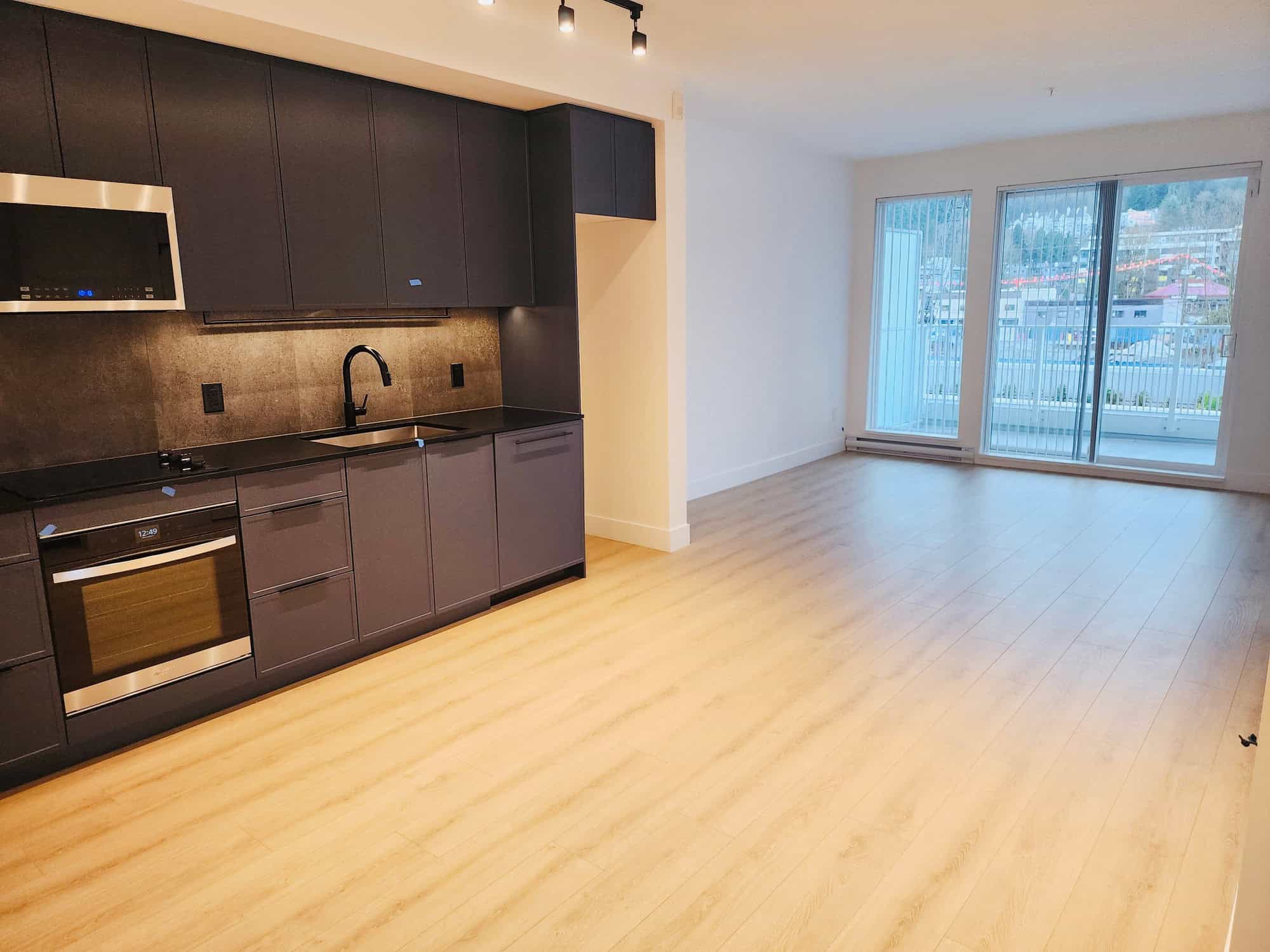Modern open-concept kitchen and living space in a Vancouver-area apartment, featuring sleek dark cabinetry, stainless steel appliances, and large sliding glass doors to a balcony with city views.