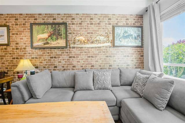 Bright living room with exposed brick wall, grey sectional sofa, and large window showcasing lush greenery, perfect for Vancouver real estate and home staging.