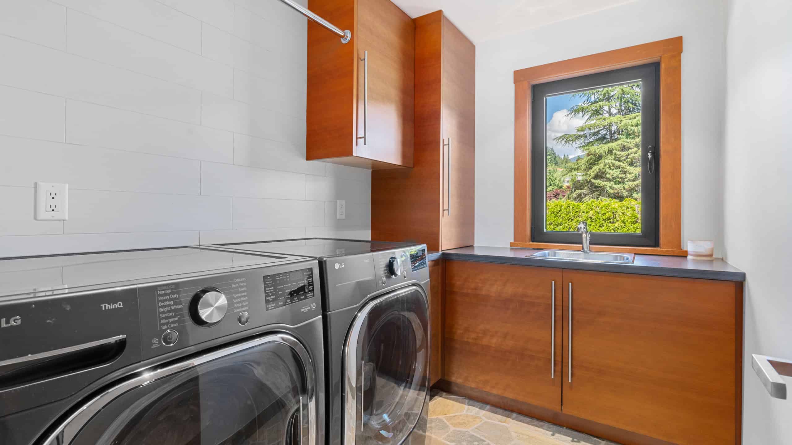 Modern Laundry Room in West Vancouver Home Bright laundry room with washer and dryer, wooden cabinets, and a large window overlooking lush greenery in West Vancouver.