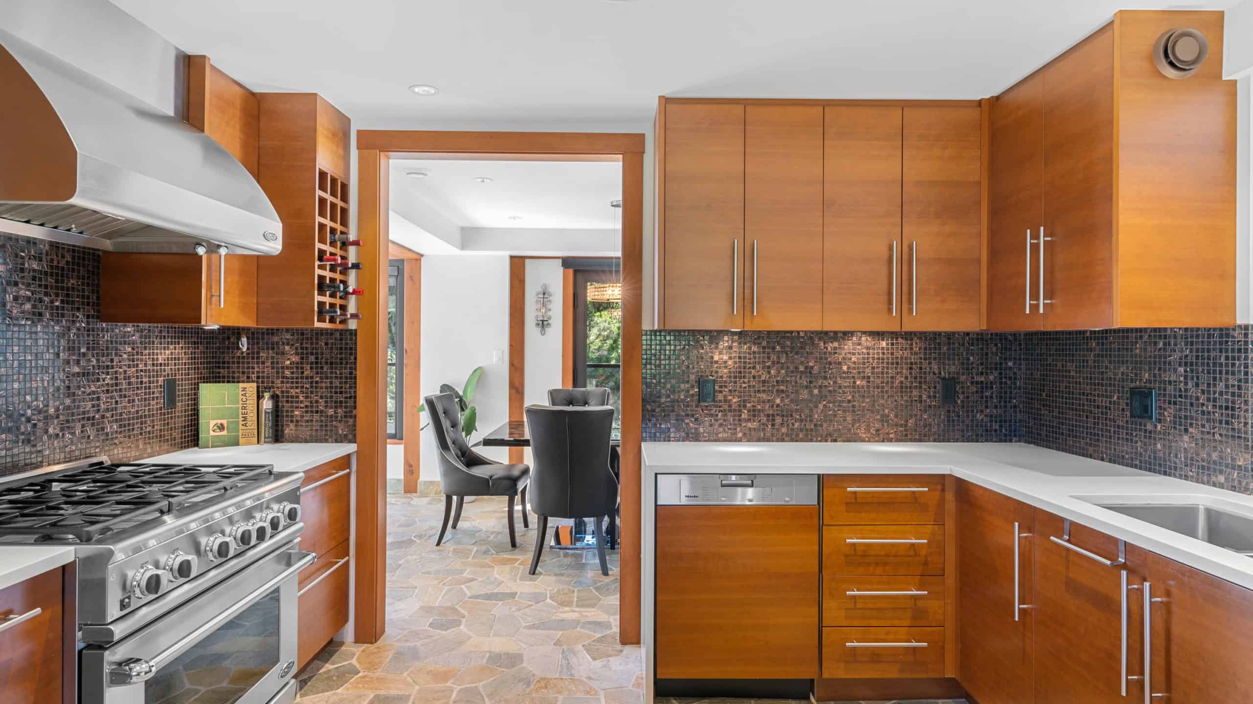 Modern Kitchen with Wooden Cabinets and Stone Floor Spacious kitchen featuring warm wooden cabinetry, a stainless steel stove, and a mosaic tile backsplash, seamlessly connecting to a dining area with natural light.