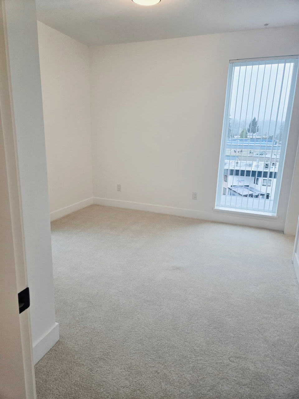 Bright, clean empty bedroom with large window and vertical blinds, featuring neutral carpet flooring and white walls, ideal for Vancouver condo or townhouse living.