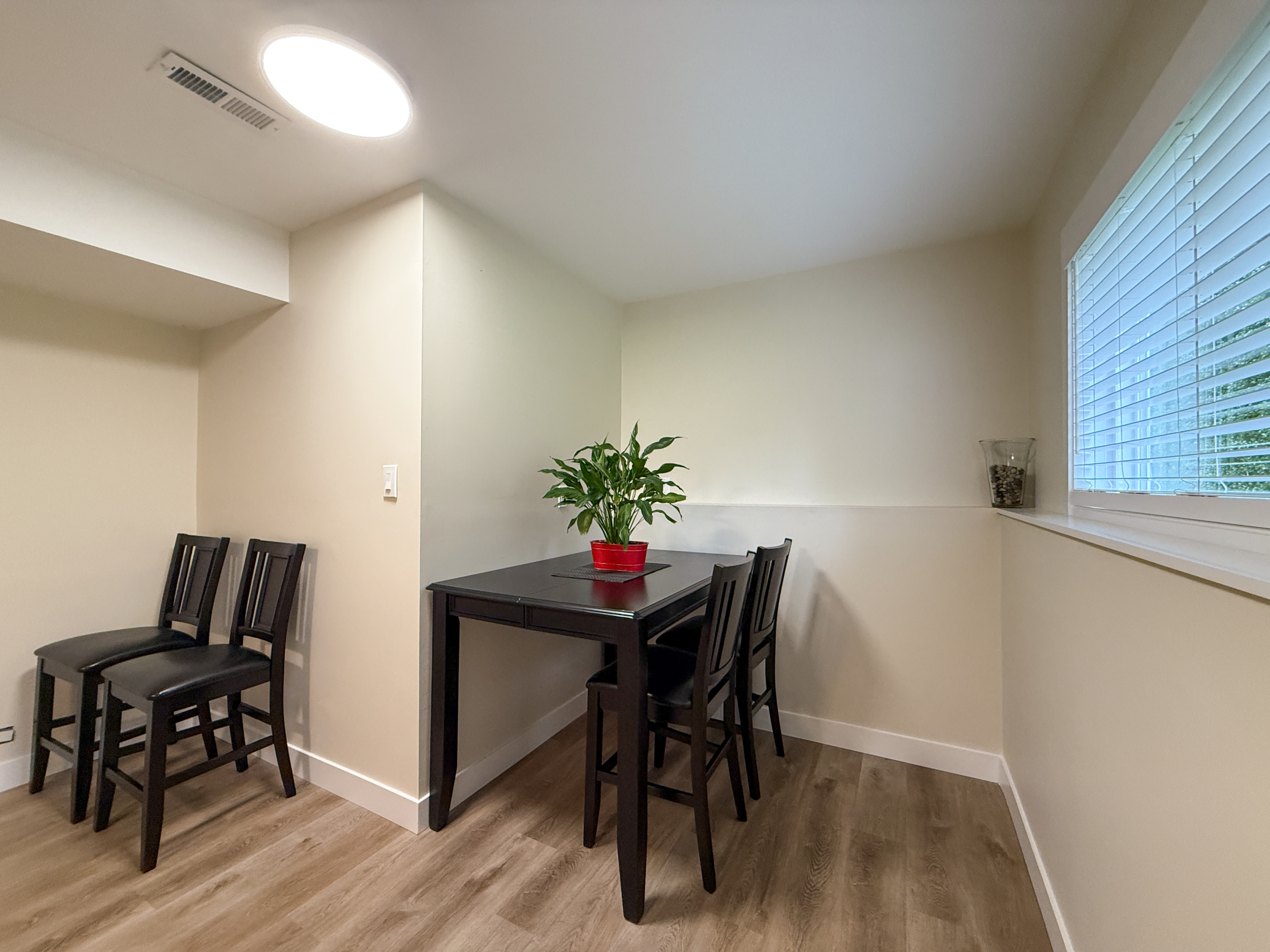 Bright interior view of a dining space in North Vancouver, featuring a black dining table with four chairs, a window with blinds, and minimal decor.