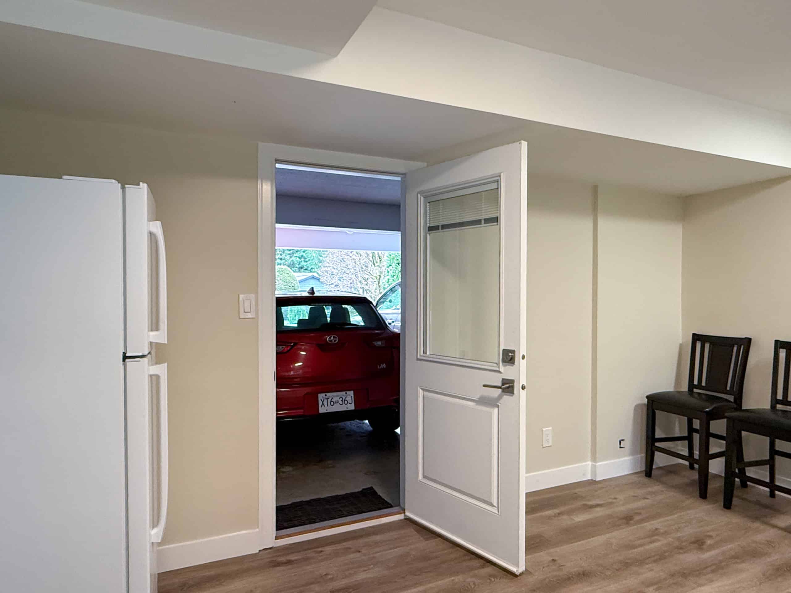 Bright interior view of a home with open door leading to garage, showcasing a red car parked outside, and minimalistic decor with chairs along the wall.