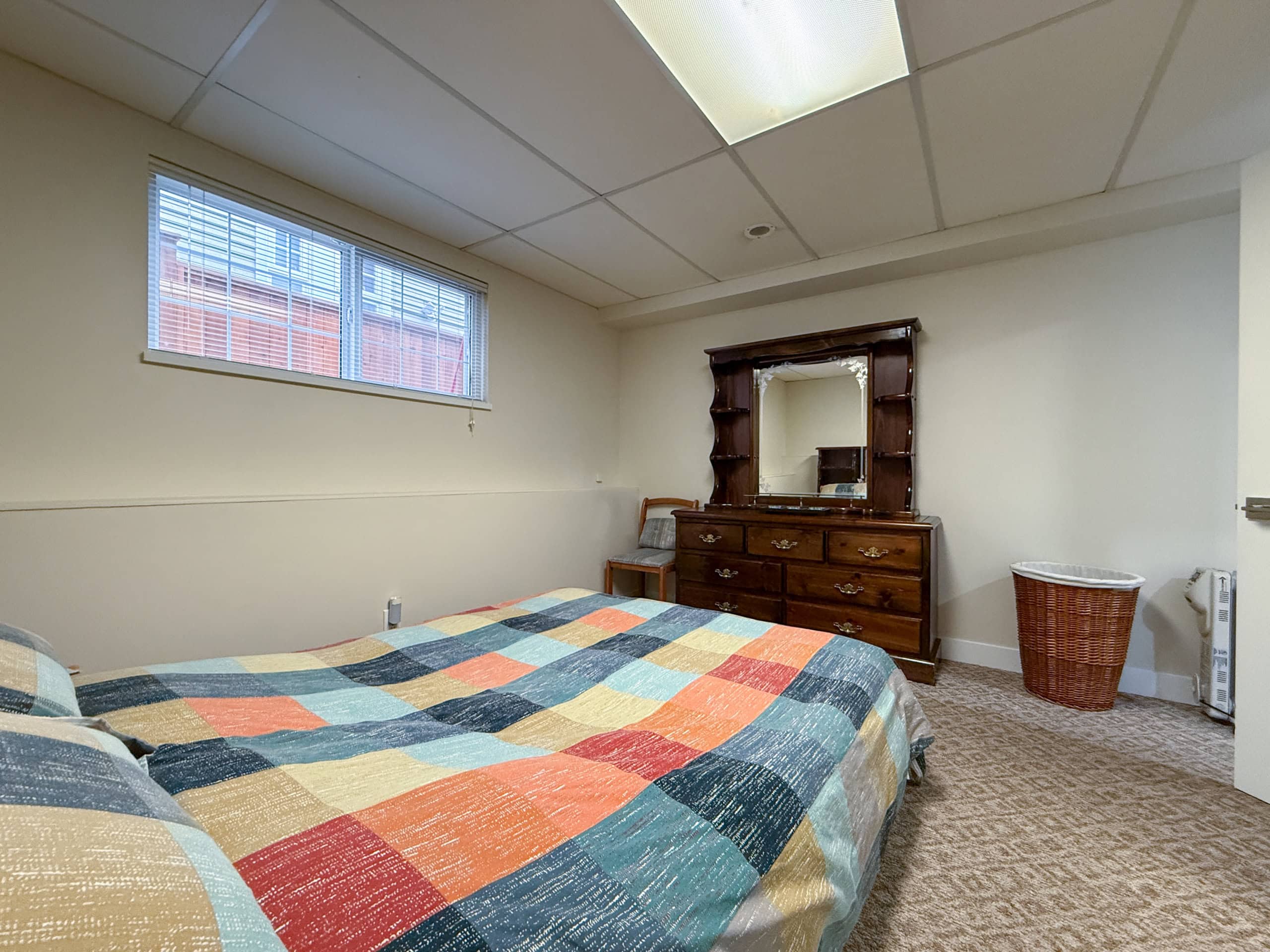 Interior view of a North Vancouver bedroom with a large window overlooking Dempsey Road, featuring a cozy bed with a colorful patchwork quilt and classic wooden furniture.