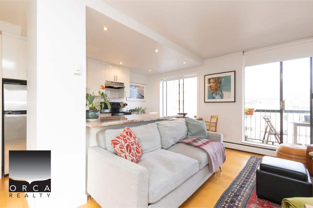 Bright, modern Vancouver condo living room with balcony, natural light, contemporary decor, and open kitchen featuring stainless steel appliances—ideal for real estate buyers.