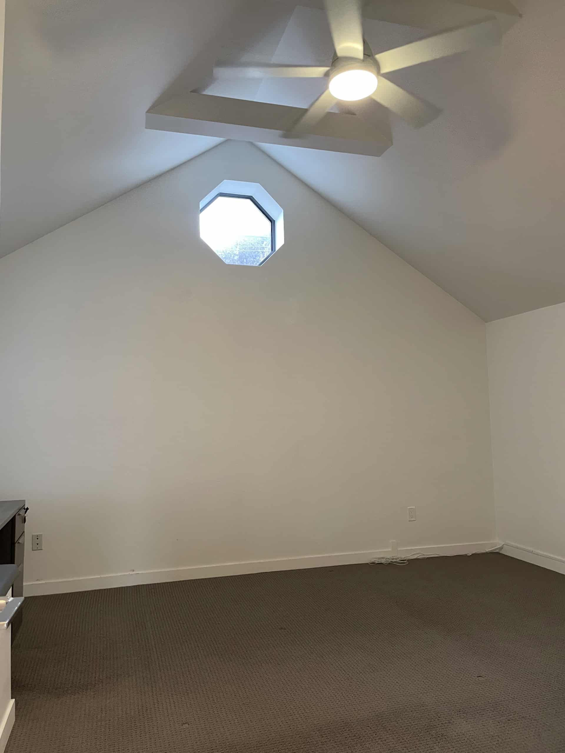 Bright empty attic room with an octagonal window, ceiling fan, and sloped ceiling, showcasing Vancouver Island real estate opportunities, perfect for renovation or conversion into a cozy living space.
