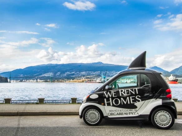 Smart black compact electric car with "We Rent Homes" advertisement, waterfront view of Vancouver, mountains, and city skyline in the background, promoting Orca Realty Inc. property rentals in BC.