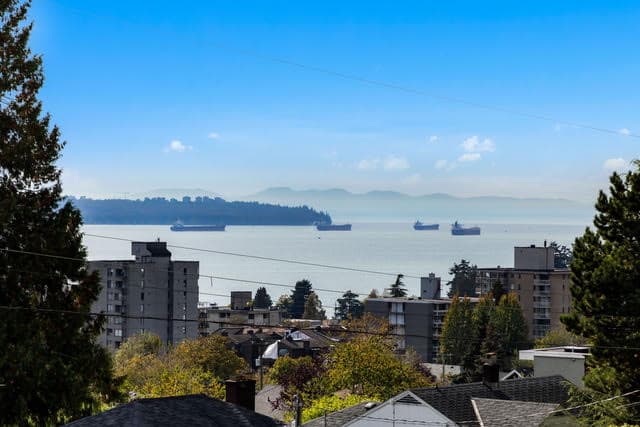 A panoramic view of West Vancouver's waterfront featuring residential buildings, lush trees, and ships on the water with mountains in the background.