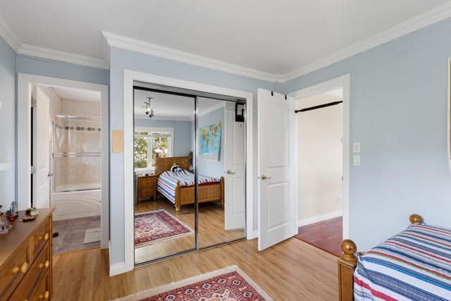 Bright and airy bedroom in a West Vancouver home with light blue walls, wooden furniture, and a mirrored closet door, showcasing a cozy and inviting space.
