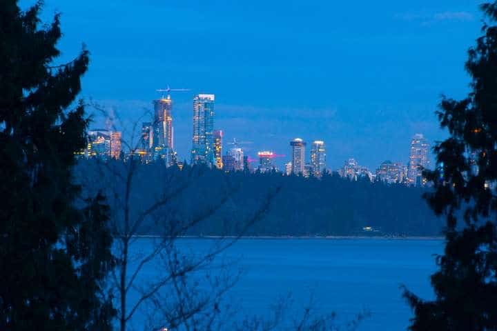 City skyline view of downtown Vancouver from West Vancouver with illuminated skyscrapers and calm water in the foreground.