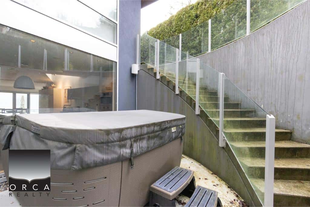 Outdoor staircase leading to a modern hot tub area with glass railing and greenery backdrop.