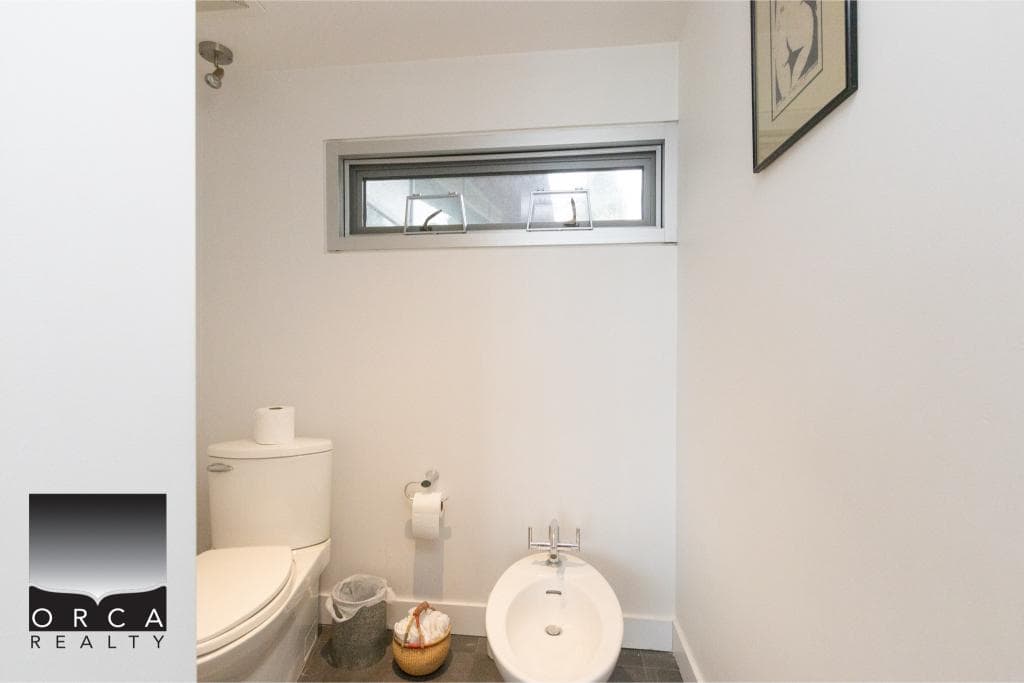 Bright, clean bathroom featuring a toilet, bidet, and simple wall art, with natural light from a narrow window.