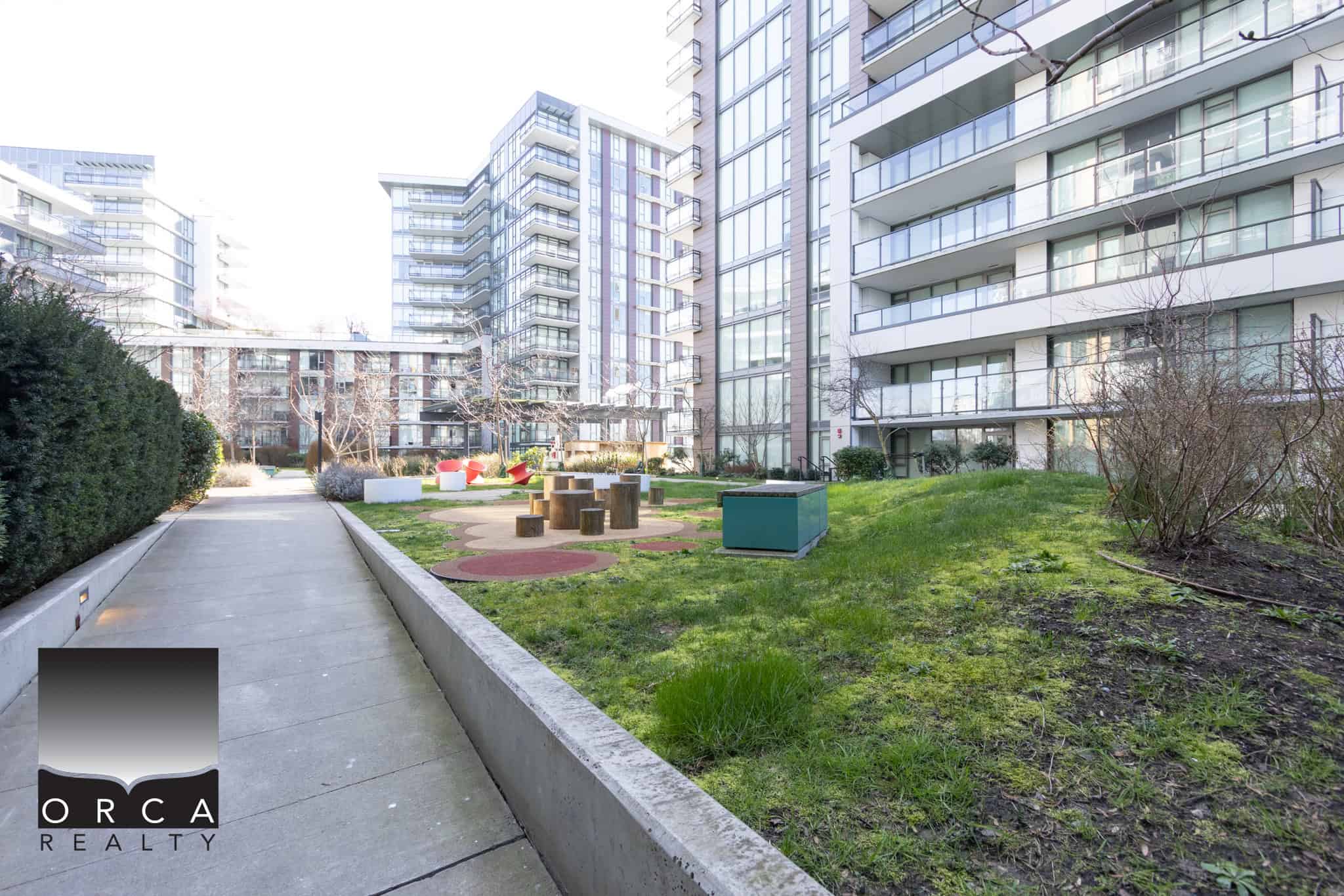 Modern Richmond apartment complex with landscaped outdoor area and seating. Spacious outdoor courtyard at 702E-3280 Corvee Way, Richmond, featuring greenery, seating, and walking paths in a contemporary residential community.