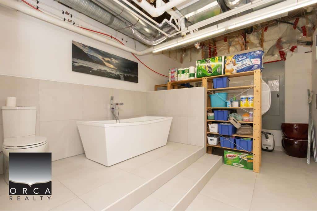 Bright laundry room featuring a sleek bathtub, organized shelving with cleaning supplies, and exposed ceiling pipes in Ansell Place.