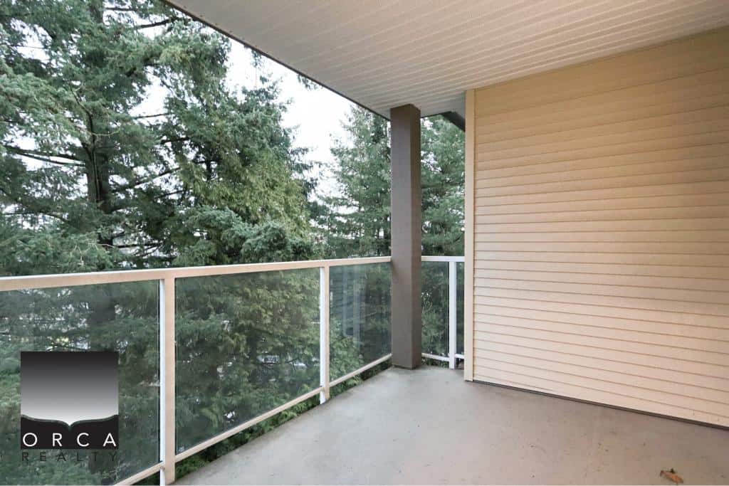 Spacious balcony at 410-15895 84th Ave, Surrey, featuring glass railings and a view of mature trees, perfect for relaxing or outdoor entertaining in a serene residential setting.