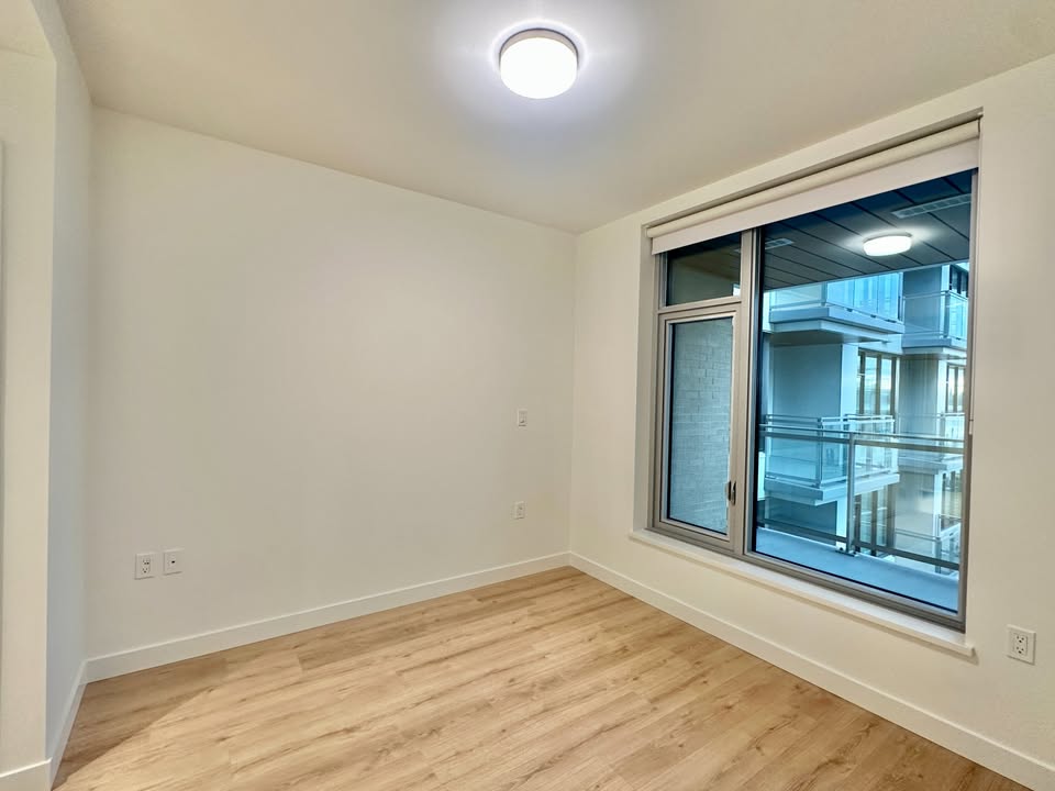 Bright, empty condo room in North Vancouver with large window and hardwood flooring, ideal for real estate listings.