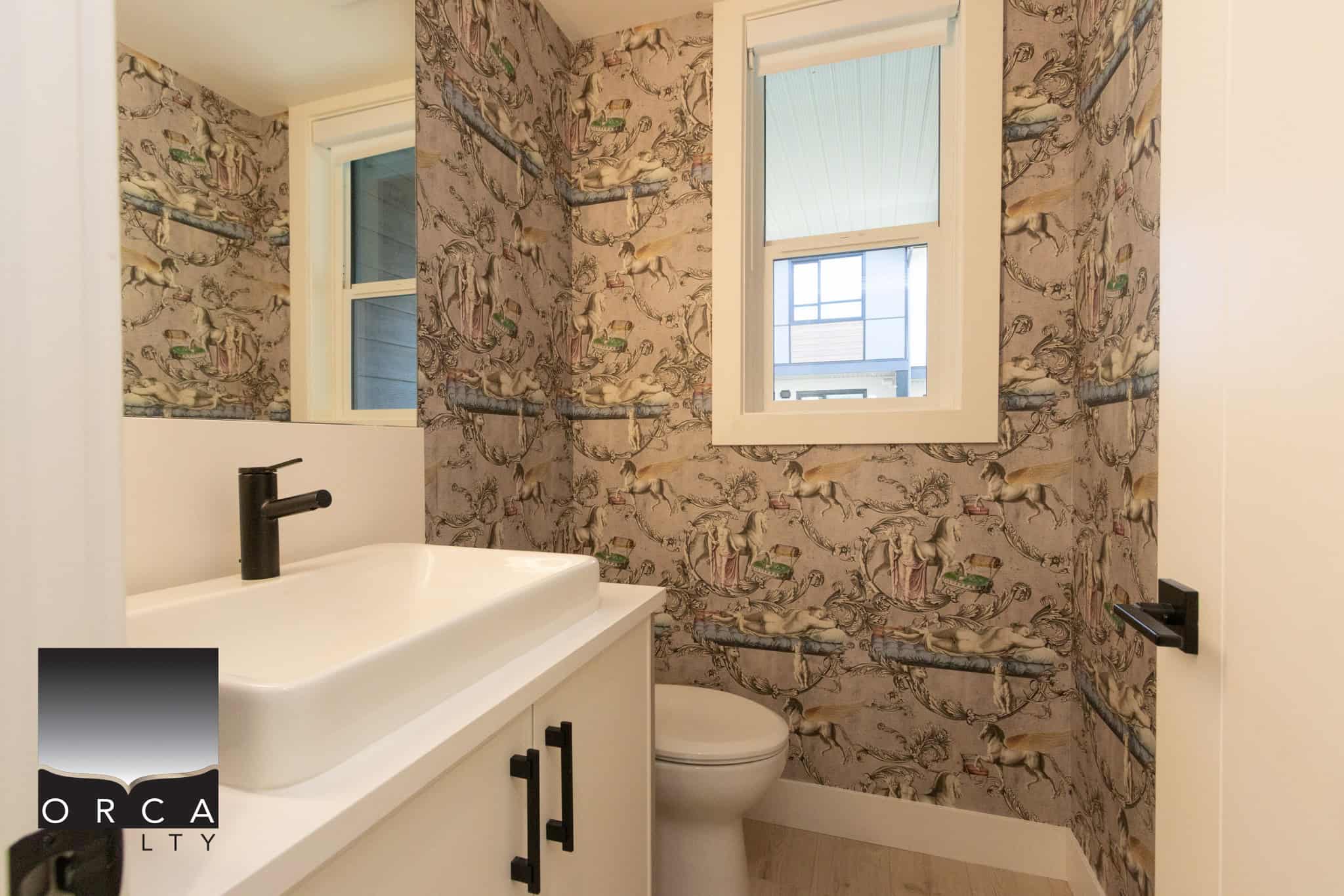 Bright bathroom featuring a modern sink, black fixtures, and a decorative wallpaper with colorful illustrations. Natural light from two windows enhances the cozy, stylish space.
