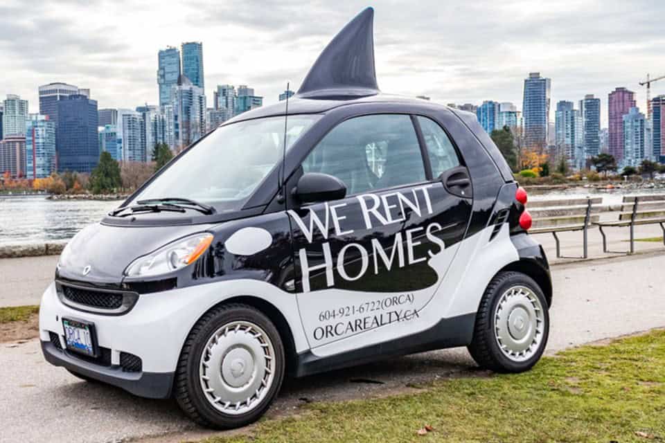 Compact rental car with "We Rent Homes" signage parked near Vancouver skyline, ideal for property tours and client visits in West Vancouver.