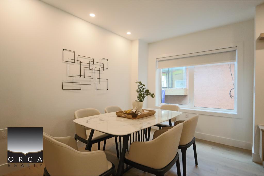 Elegant dining area featuring a white marble table, six beige upholstered chairs, and contemporary wall art, illuminated by recessed ceiling lights and a large window with a view of the outdoors.