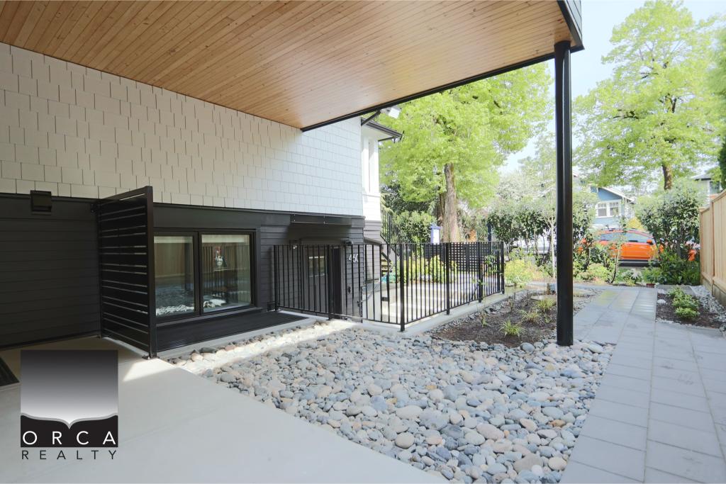 Contemporary backyard patio with garden and stone pathway, featuring a black metal fence and lush green trees, ideal for outdoor living and relaxation.