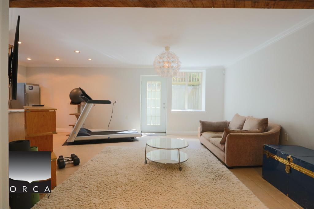 Bright and spacious living room featuring a treadmill, sofa, and large window, ideal for home fitness and relaxation. Perfect for showcasing residential real estate in Victoria, BC.