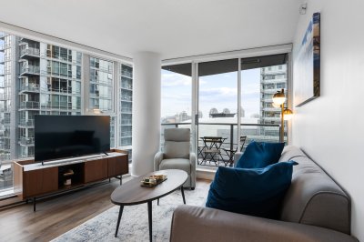 A modern living room with large windows offering city views, featuring a sleek TV stand, comfortable sofa with blue cushions, and a cozy armchair, ideal for urban residential living in Vancouver.