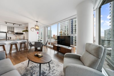 A comfortable, modern living room in a high-rise apartment with large windows offering city views, featuring contemporary furniture, hardwood floors, and an open-concept design.