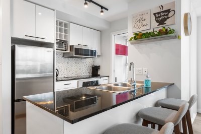 Modern white kitchen with stainless steel appliances, granite countertops, and a breakfast bar with seating, featuring bright natural light and contemporary decor.
