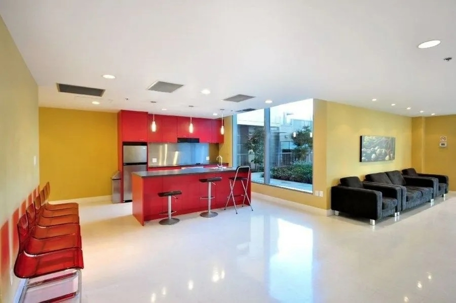 Bright modern condo interior with a colorful kitchen featuring red cabinets, a breakfast bar with stools, and a spacious living area with black sofas and large windows offering natural light.