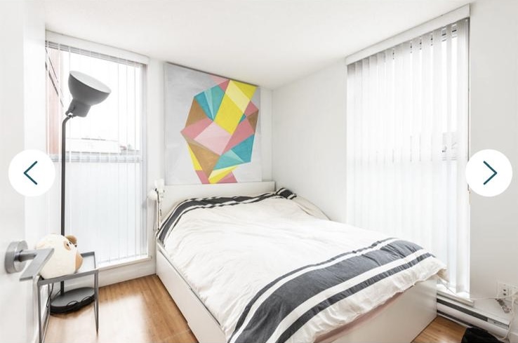 Bright modern bedroom featuring a white bed, colorful geometric wall art, and large windows with vertical blinds for natural light.