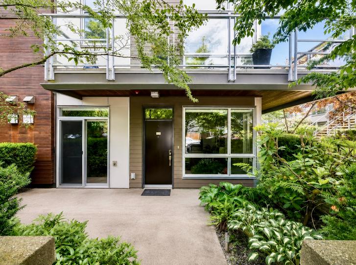 Contemporary North Vancouver home entrance with large glass windows, lush greenery, and a balcony above, showcasing modern architecture and outdoor landscaping.