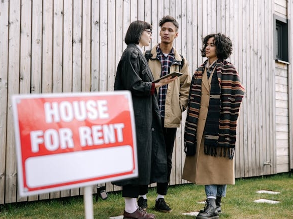 Landlord and two tenants stand outside a wooden house with a House for Rent sign discussing rental details