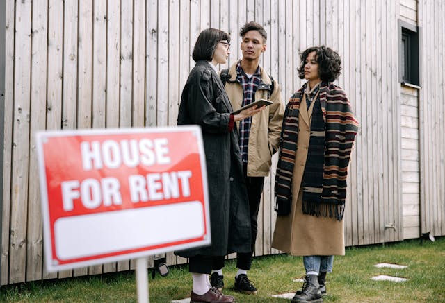 Landlord and two tenants stand outside a wooden house with a House for Rent sign discussing rental details