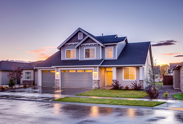 Modern two-story suburban house with gray siding, dark roof, and warm interior lights reflecting on wet pavement